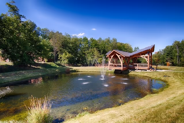 A serene outdoor scene featuring a small pond with a water fountain in the center, surrounded by green grass and trees. There is a wooden gazebo with a peaked roof extending over part of the pond, and a clear blue sky overhead.