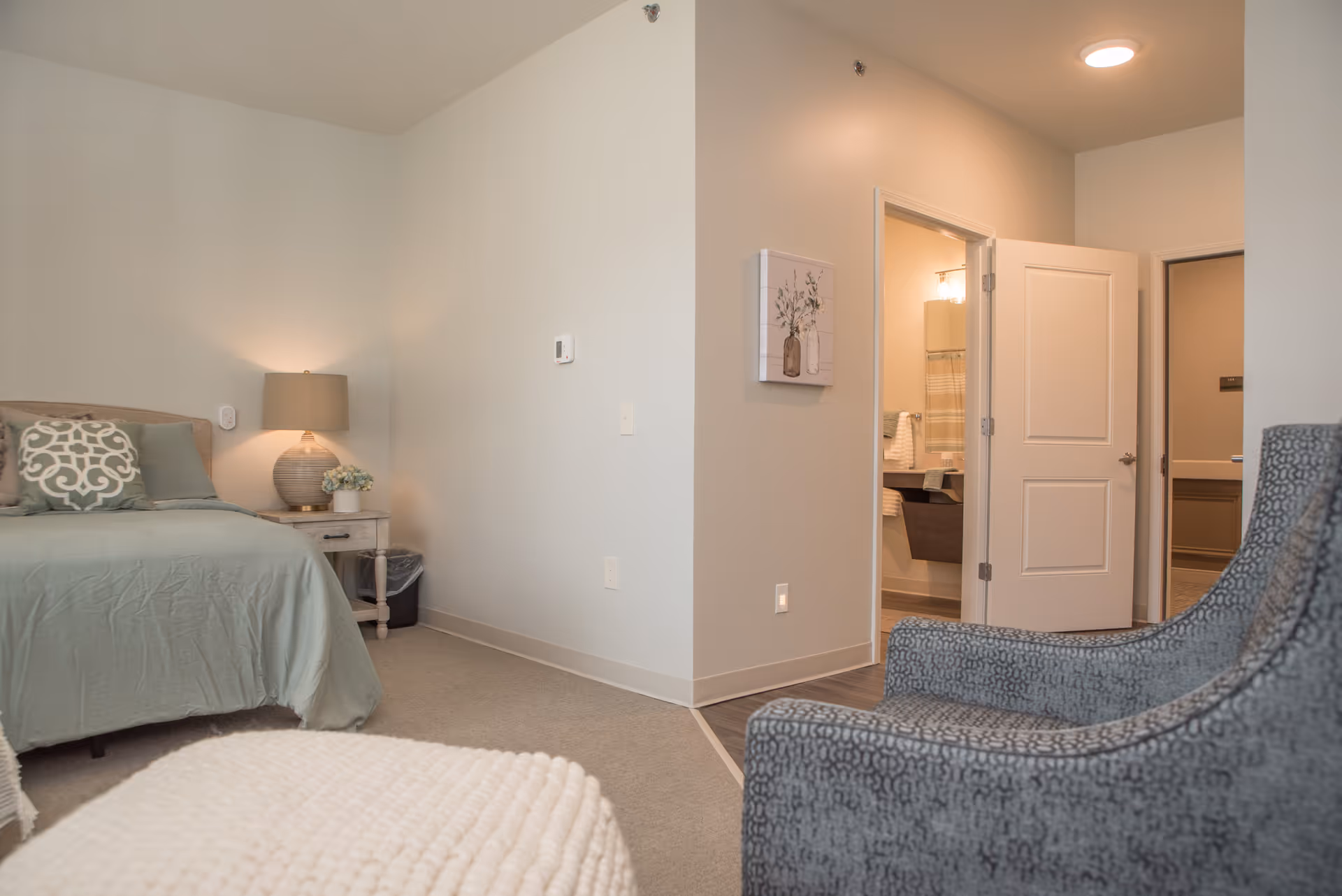A cozy senior living bedroom with a bed covered in green bedding and decorative pillows, a bedside table with a lamp and small plant, a gray patterned armchair, and an open door leading to a bathroom with towels and a mirror visible.
