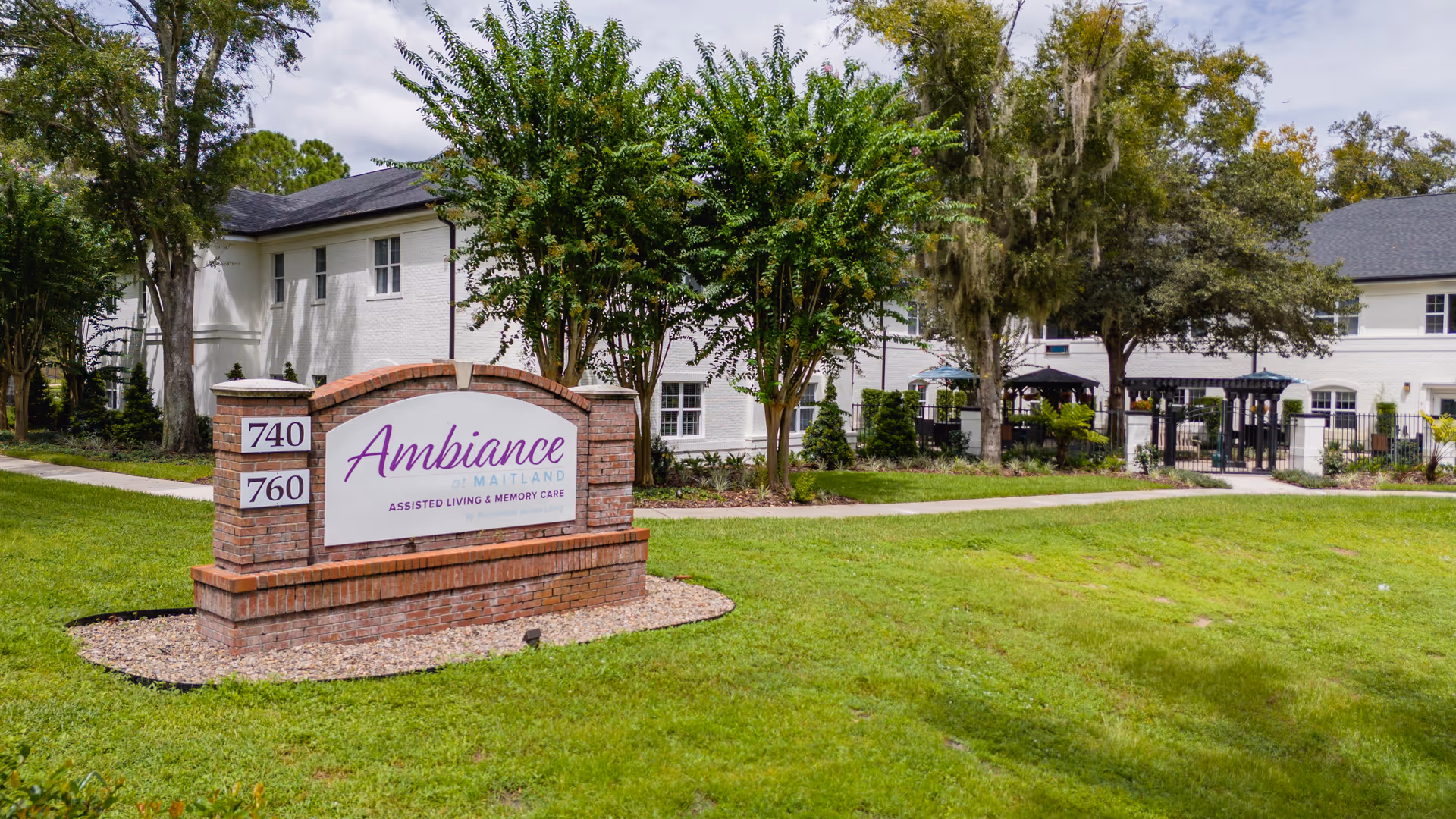 Exterior view of Ambiance at Maitland assisted living and memory care facility with a brick sign displaying the name and address numbers 740 and 760, surrounded by green grass, trees, and a white building in the background.