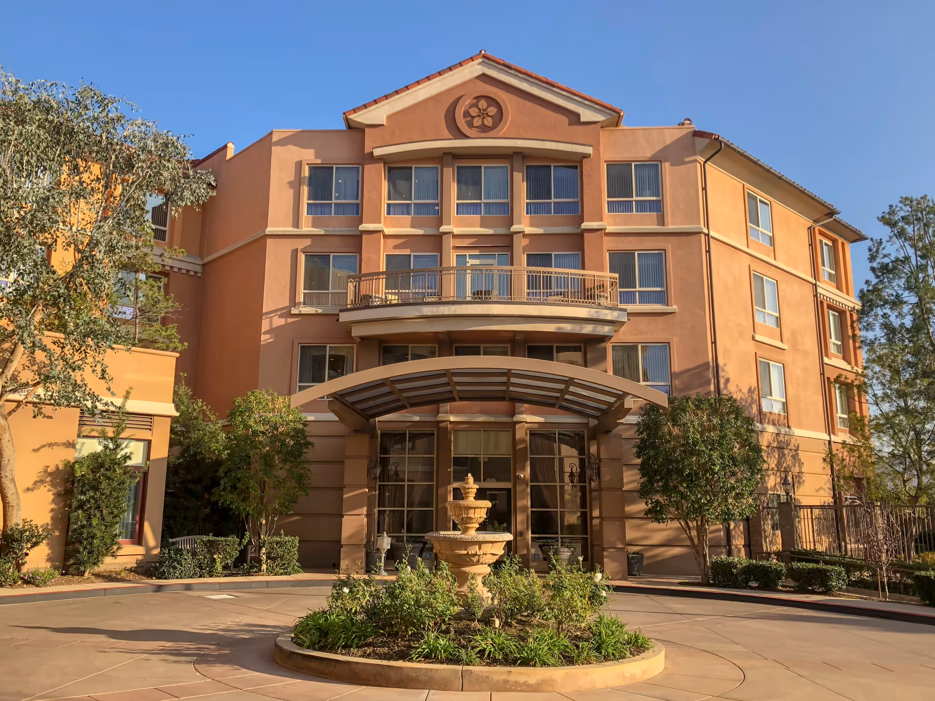 Front exterior view of a multi-story senior living facility building with a covered entrance, a circular driveway, a central fountain, and landscaped greenery under a clear blue sky.