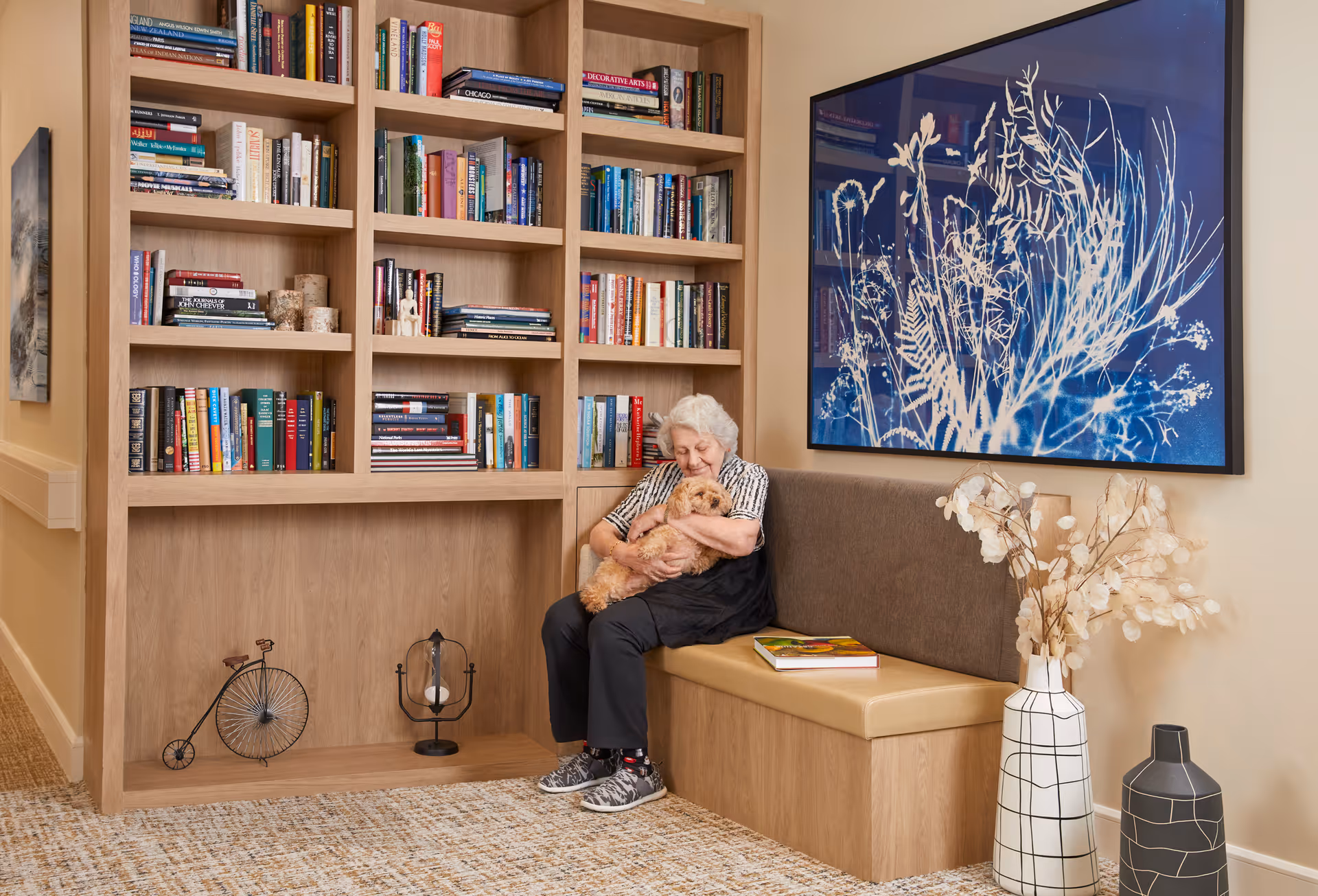 An elderly woman sitting on a cushioned bench in a cozy reading nook, holding a small dog. Behind her is a large wooden bookshelf filled with books and decorative items. On the wall above the bench is a large blue and white botanical artwork. Two decorative vases with dried plants are placed on the floor nearby.