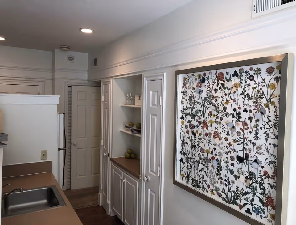 Small galley-style kitchen with a sink and countertops, built-in cabinets and shelves, and a large framed floral print on the wall.