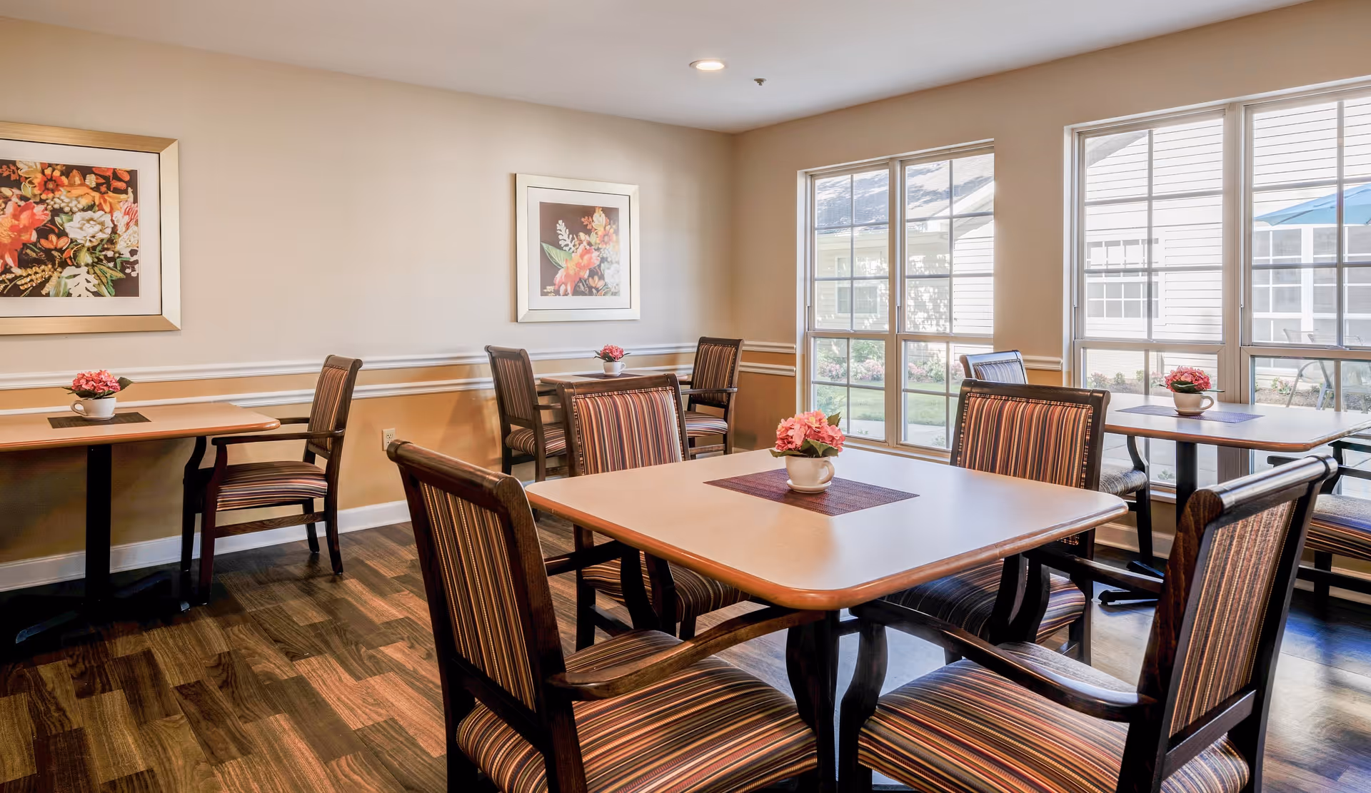 A bright dining room with several wooden tables and striped cushioned chairs. Each table has a small flower arrangement in a white pot. The room has large windows letting in natural light and two framed floral paintings on the beige walls. The floor is dark wood.