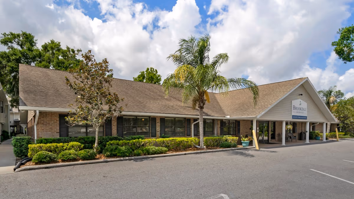 Exterior view of Brookdale New Port Richey senior living facility showing a single-story building with a sloped roof, large windows, palm trees, and landscaped bushes in front under a partly cloudy sky.