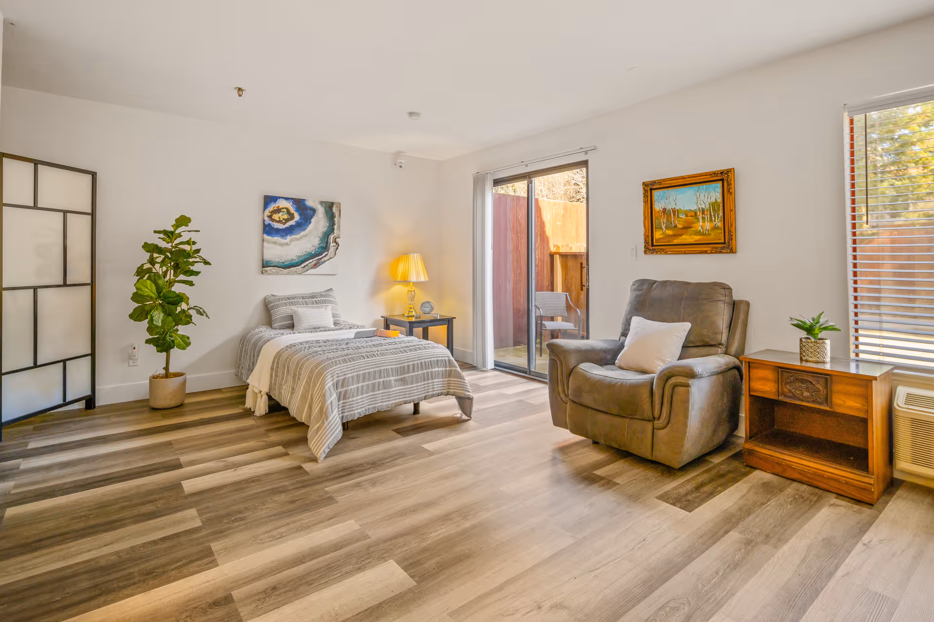 A bright and spacious bedroom with a single bed covered in striped bedding, a bedside table with a lamp, a large armchair with a pillow, a wooden nightstand with a small plant, a potted plant, a folding room divider, and two paintings on the walls. There is a sliding glass door leading to an outdoor patio and a window with blinds letting in natural light.