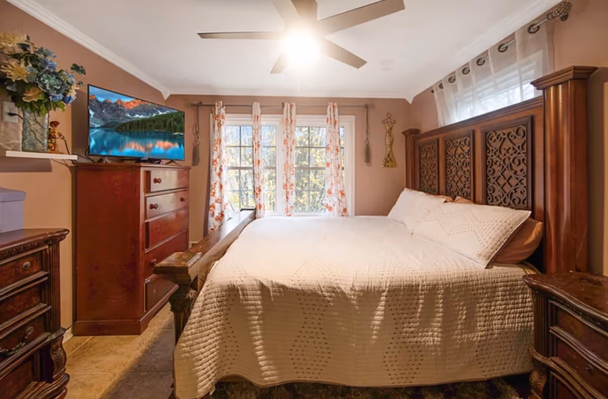 Cozy bedroom with an ornate wooden headboard, a quilted bed, dresser with a TV, ceiling fan, and a window with floral curtains.