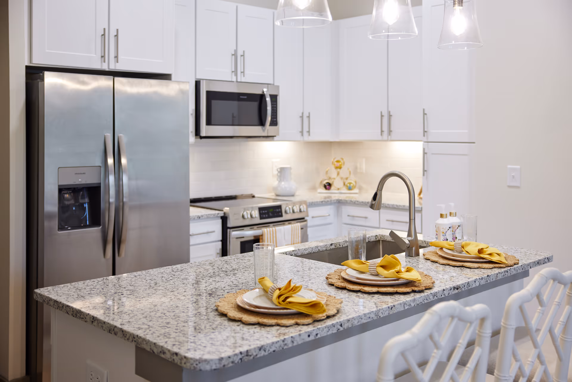 Modern white kitchen with a granite island set with place settings, stainless steel appliances, and pendant lights.
