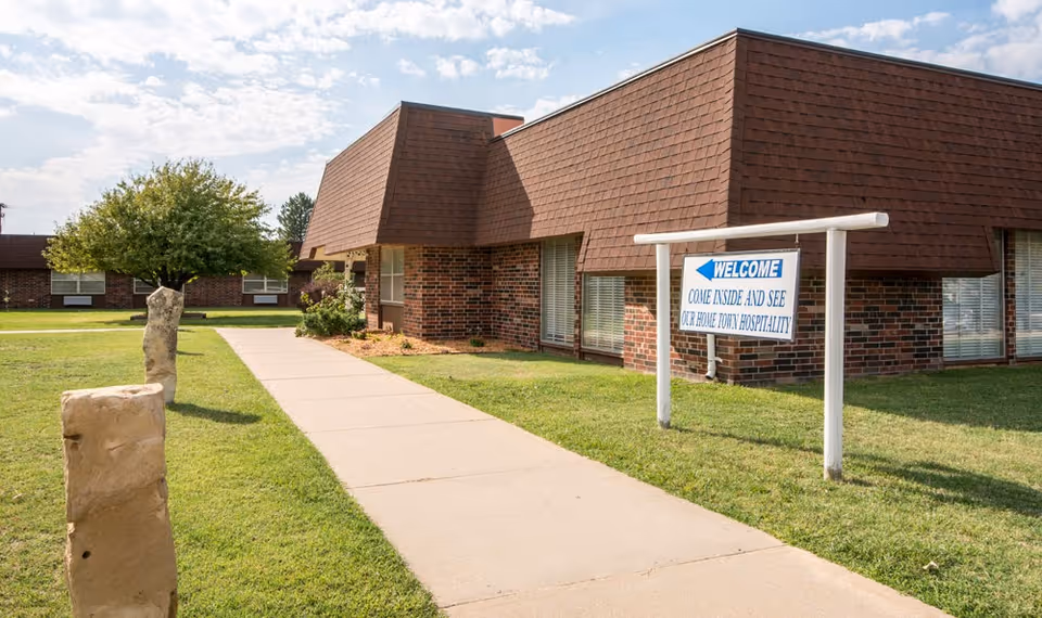 Exterior view of a brick building with a brown shingled roof and large windows. A concrete walkway leads up to the building, flanked by grass and stone pillars. A white sign with blue text reads 'WELCOME COME INSIDE AND SEE OUR HOME TOWN HOSPITALITY.' Trees and another building are visible in the background under a partly cloudy sky.