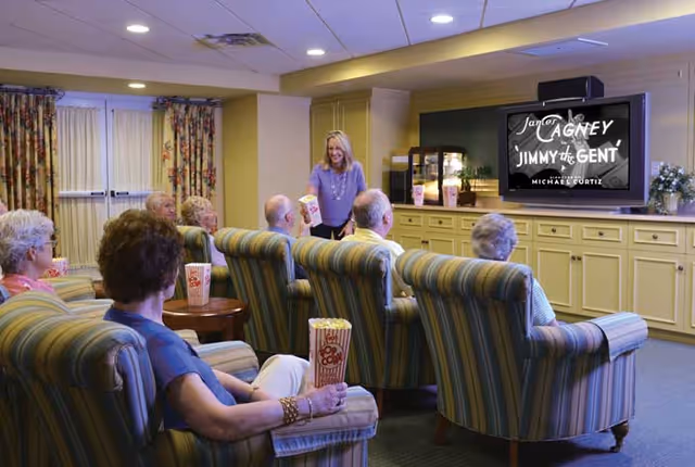 Seniors sit in striped armchairs in a communal media room eating popcorn while watching a movie on a large TV.