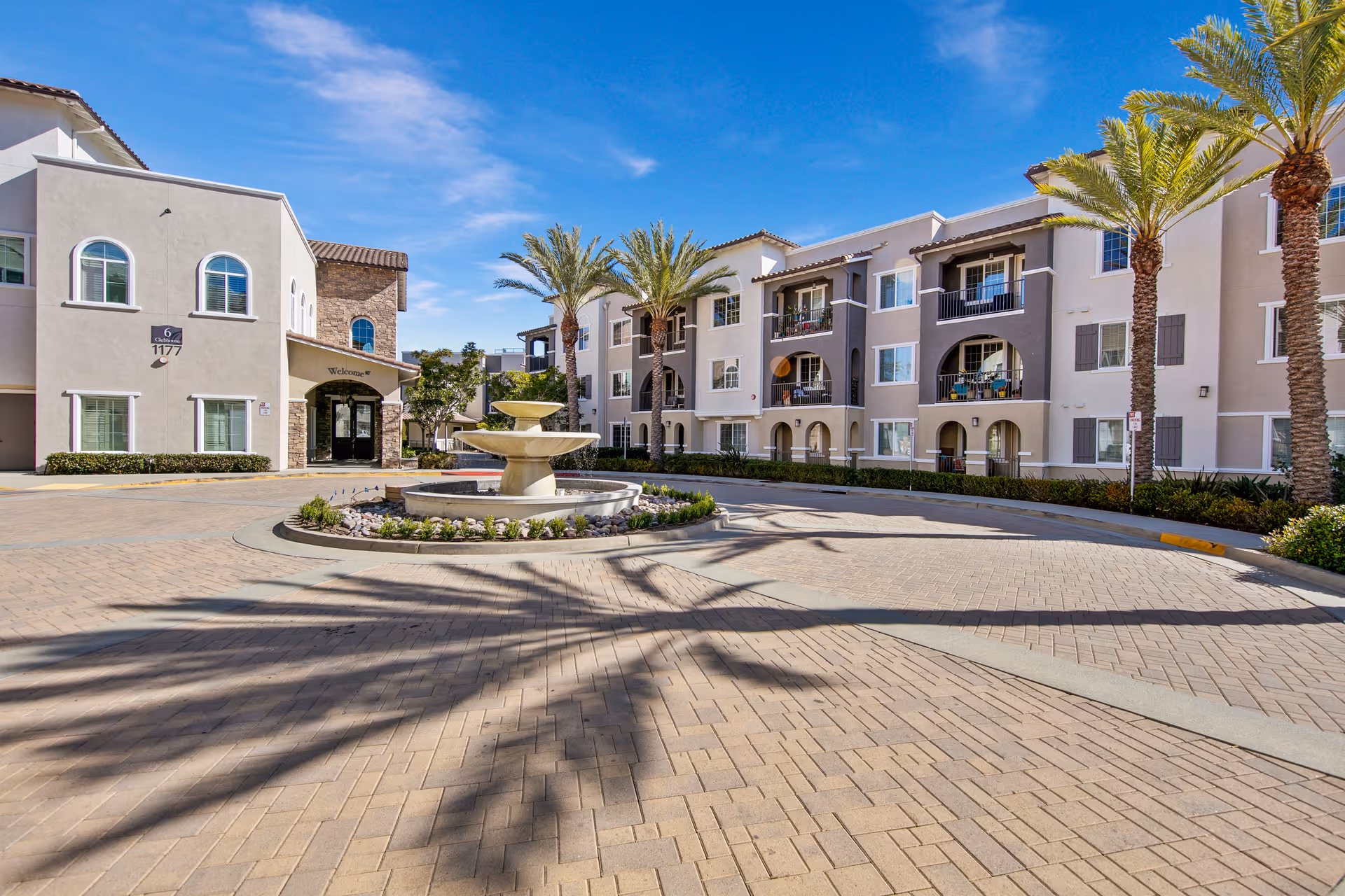 Driveway and fountain in front of a three-story Mediterranean-style senior living building with balconies and palm trees.