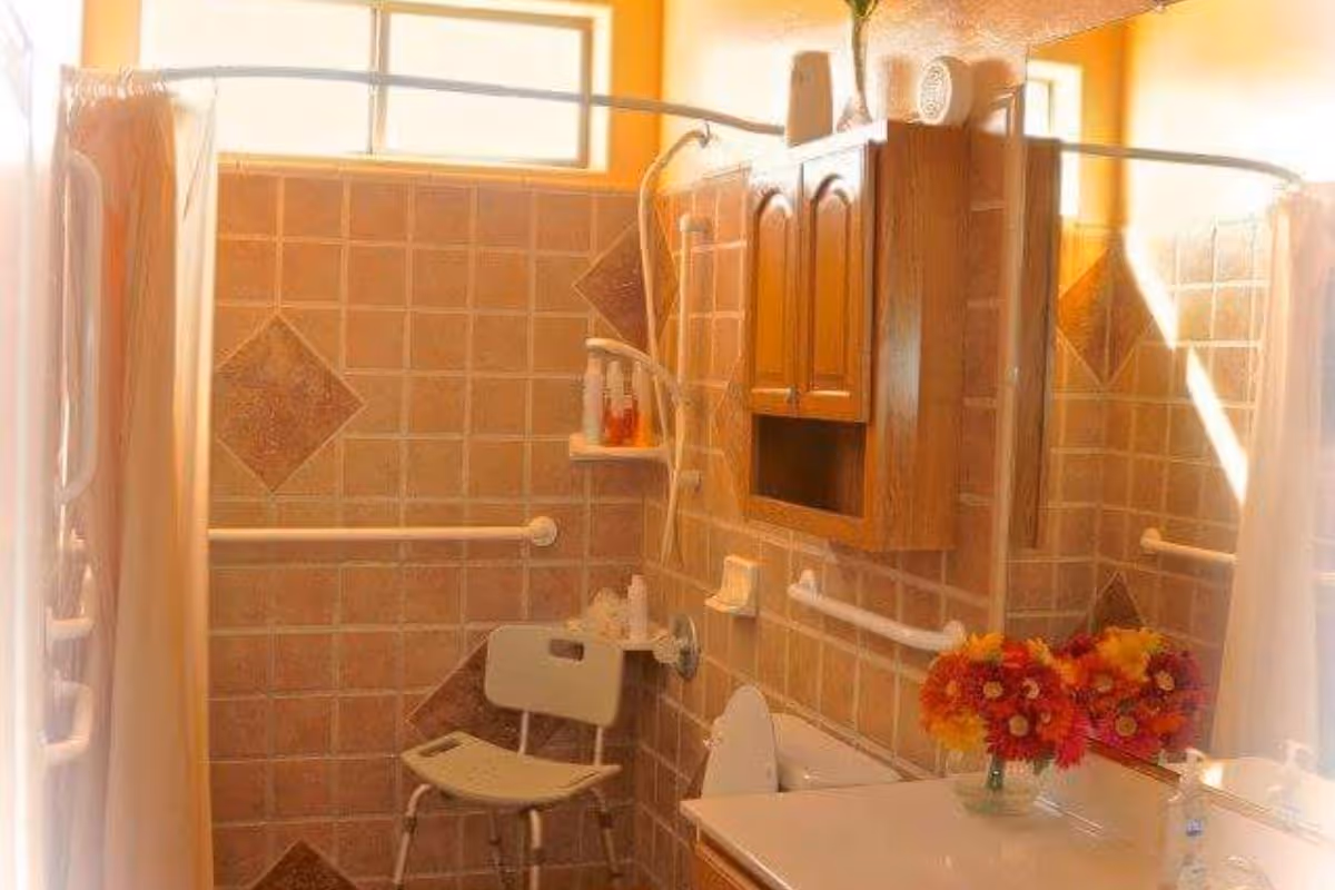 A bathroom with beige and brown tiled walls, a shower area with a white shower chair, grab bars, a wooden cabinet mounted on the wall, and a countertop with a vase of colorful flowers and a soap dispenser.