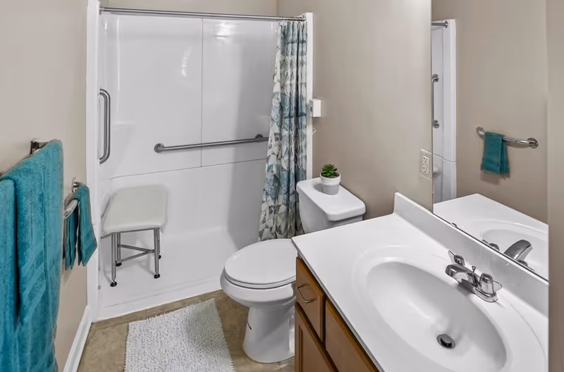 A clean and accessible bathroom featuring a white toilet with a small potted plant on the tank, a white sink with a silver faucet set in a wooden vanity, a large mirror above the sink, a white shower with a grab bar and a foldable shower seat, a blue patterned shower curtain, and teal towels hanging on the wall. The floor has a white bath mat and beige tiles.