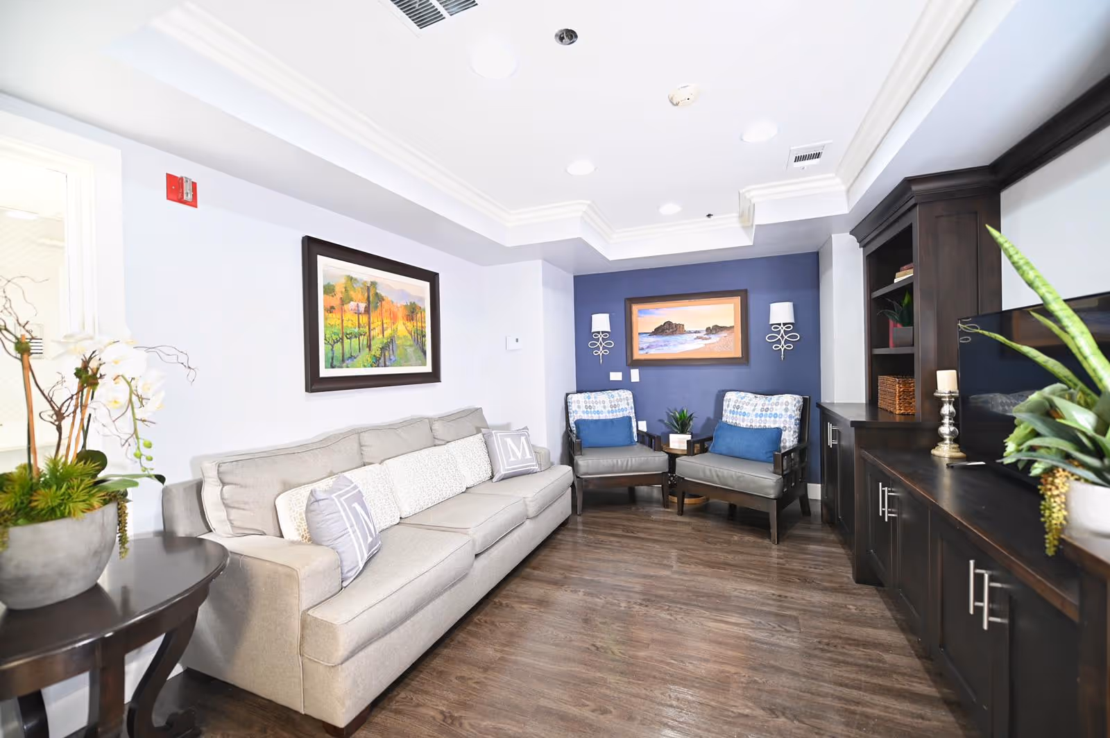 Well-lit sitting area with a beige sofa, two upholstered chairs, framed artwork, and dark wood cabinets.