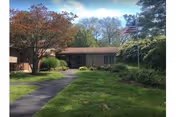 A single-story brick facility front with a walkway, lawn, trees, and an American flag.