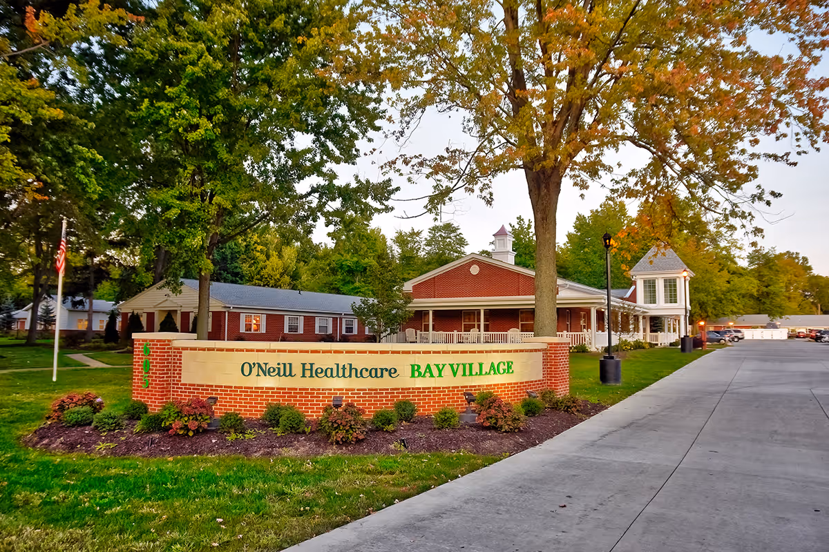 Exterior view of O'Neill Healthcare Bay Village facility with a brick sign in the foreground surrounded by landscaping. The building is a single-story structure with a porch and a small tower-like feature. Trees with green and autumn-colored leaves are visible around the property, and a paved driveway runs alongside the building.