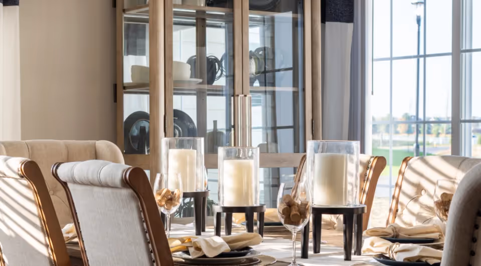 Sunlit dining room table set with large glass candle holders, upholstered chairs, and a china cabinet by tall windows.