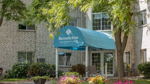 Entrance to Benedictine Living Community-Anoka featuring a blue canopy with the facility's name, surrounded by greenery, flowers, and outdoor seating in front of a brick building.