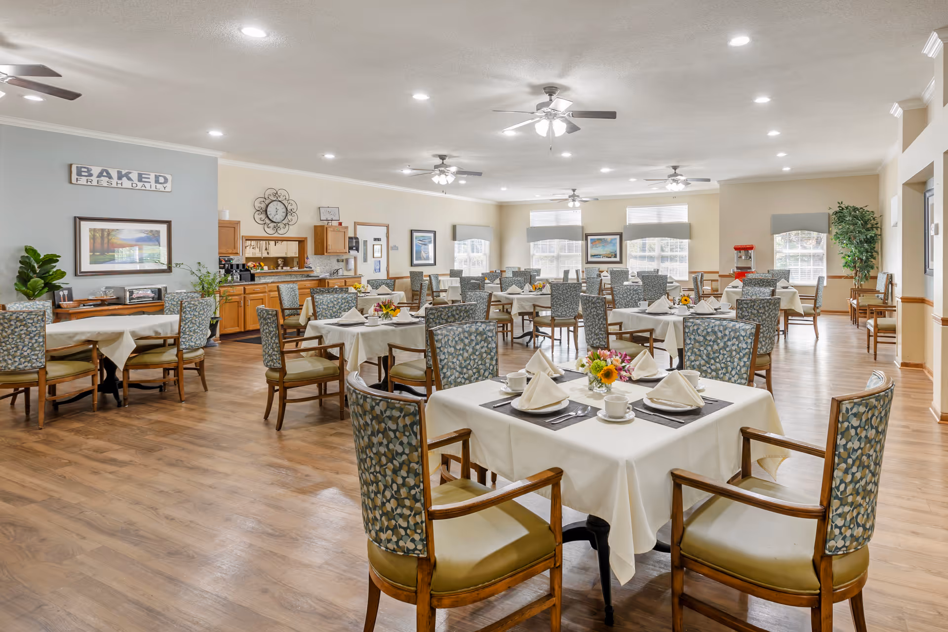 A spacious dining room with multiple tables covered in white tablecloths, each set with napkins, cups, and small flower arrangements. The room has wooden flooring, ceiling fans with lights, large windows with blinds, and decorative wall art. There is a counter area with cabinets and a sign that reads 'BAKED FRESH DAILY'.