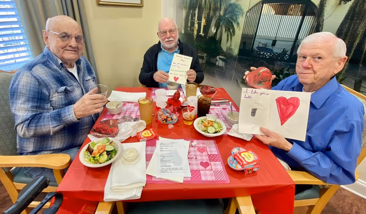 Three elderly men sitting around a table with a red tablecloth, enjoying a meal with salads, drinks, and Valentine-themed decorations. Two of the men are holding handmade Valentine cards with hearts and messages. The setting appears to be a dining area in a senior living facility.