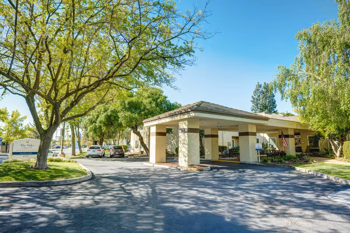 Front entrance and porte-cochère of The Oaks at Inglewood with a driveway, trees, parked cars, and a signage board.
