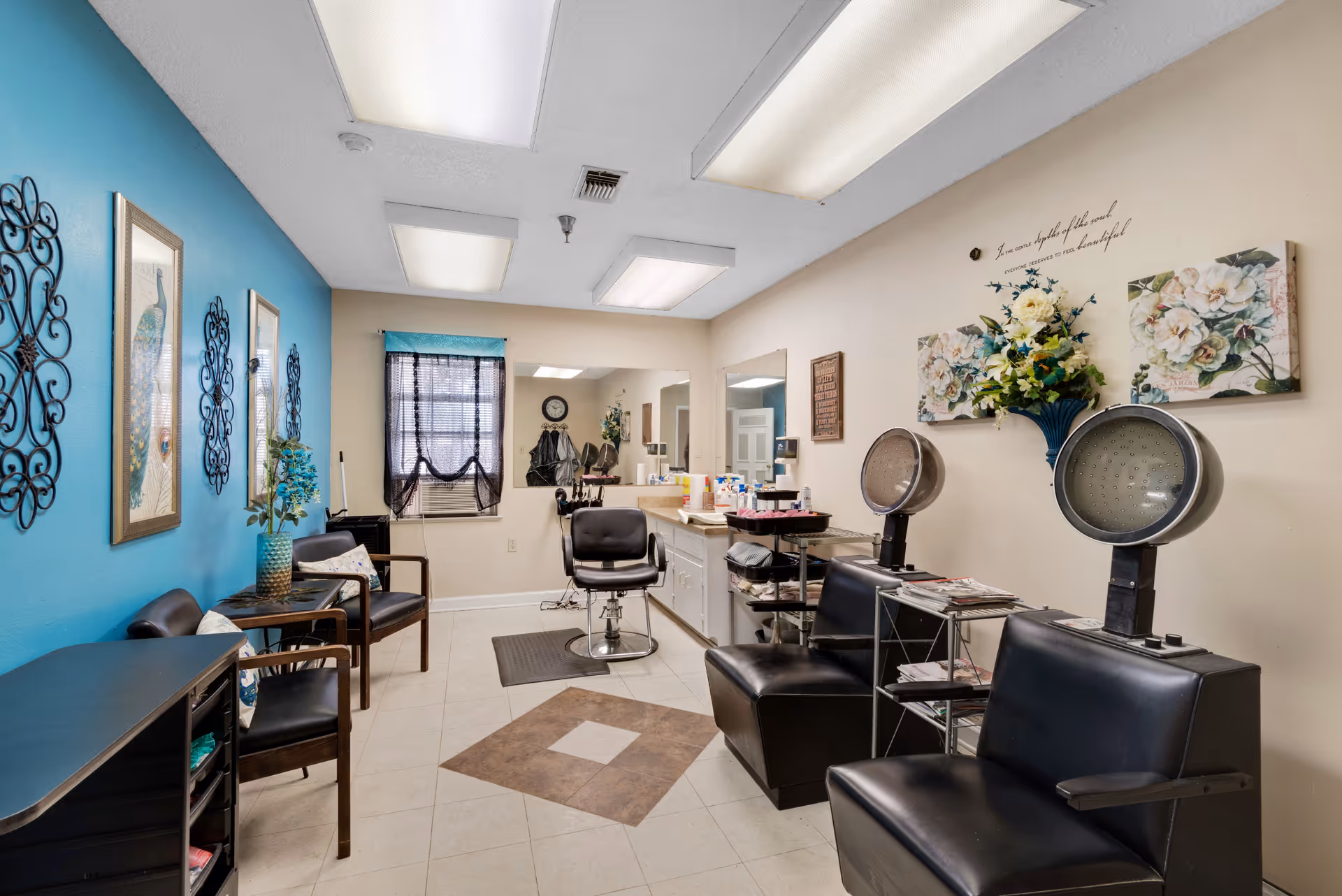 Interior of a salon room in a retirement center with two black hair drying chairs on the right, a styling chair in front of a large mirror, and seating area with chairs and decorative wall art on the left. The walls are painted beige and blue, with floral decorations and framed artwork.