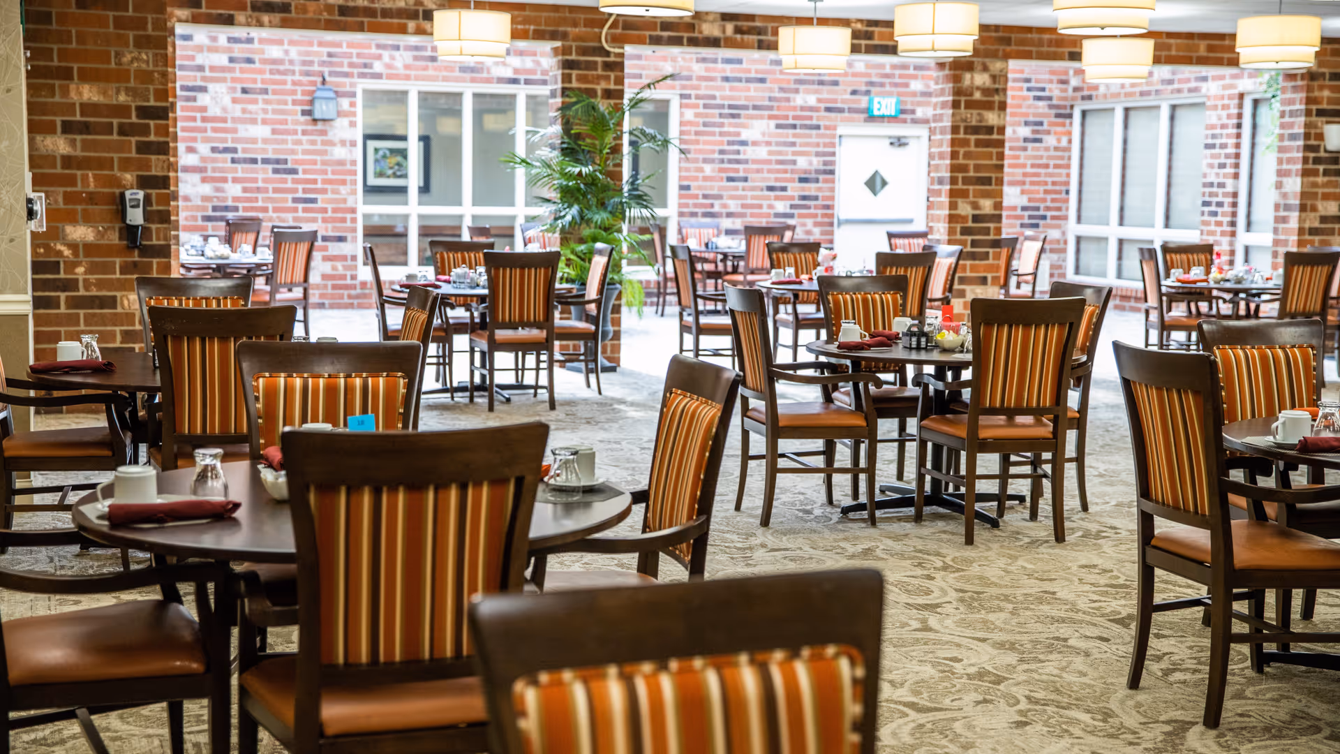 Dining room with multiple round tables and wooden chairs with striped cushions. Tables are set with cups, glasses, and napkins. The room has brick walls, large windows, and hanging ceiling lights. There is a green plant near the center and an exit door in the background.