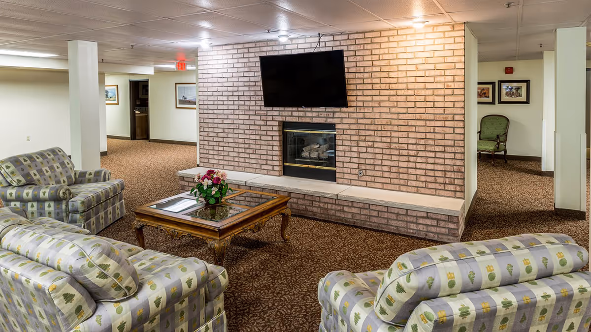 A cozy living room area with patterned upholstered sofas and armchairs arranged around a wooden coffee table with a floral centerpiece. A brick fireplace with a mounted flat-screen TV above it is the focal point of the room. The space has carpeted floors and neutral-colored walls with framed pictures hanging. There are doorways leading to other rooms in the background.