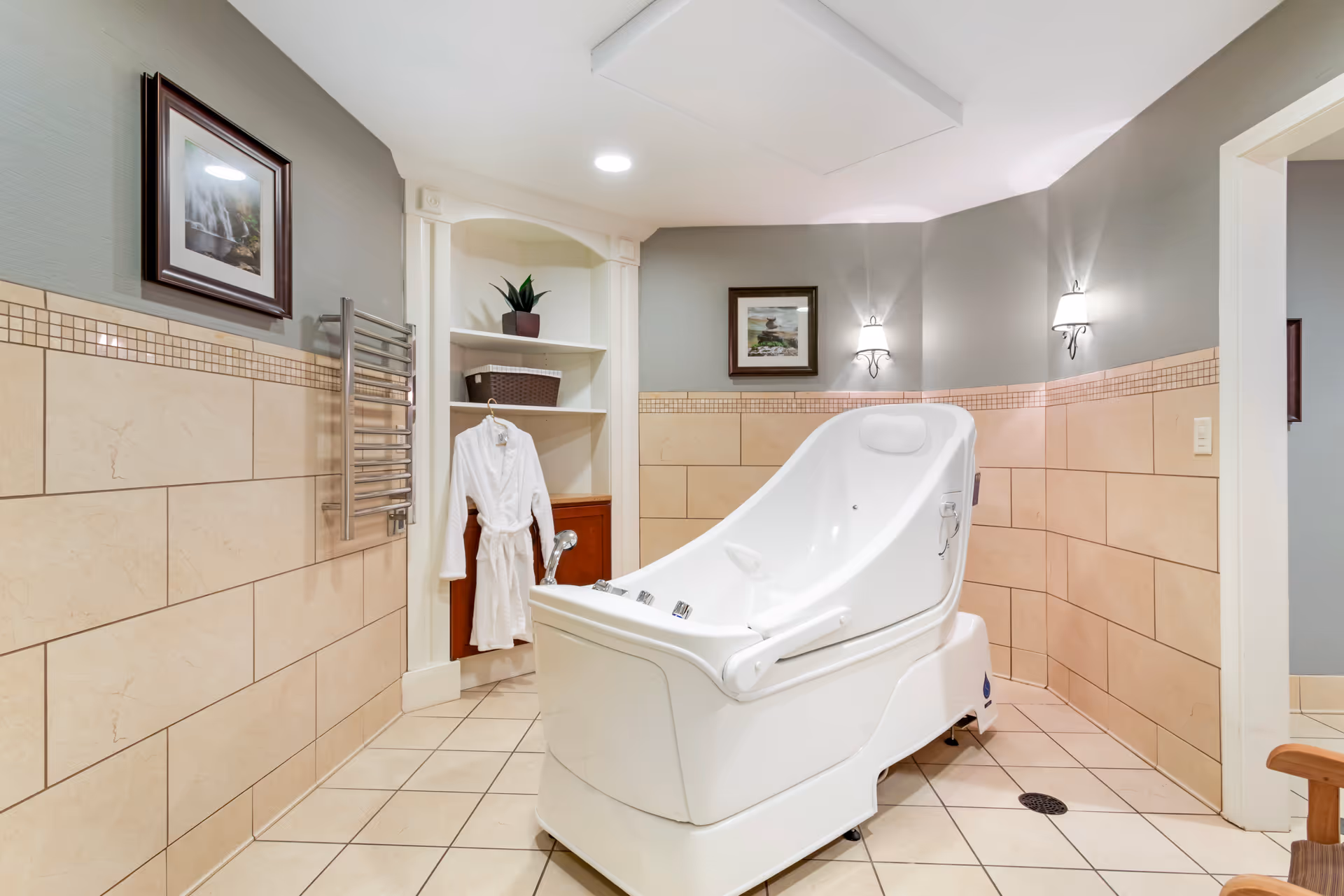 A clean, well-lit bathroom with beige tiled walls and floor, featuring a large white therapeutic bath tub with controls on the side. There is a white bathrobe hanging on a shelf unit, a towel warmer rack on the wall, two framed pictures, and two wall-mounted lights providing soft illumination.