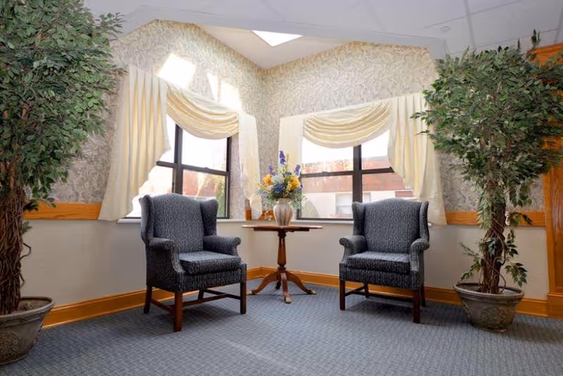 A cozy sitting area with two upholstered armchairs facing each other, separated by a small wooden table with a vase of flowers. The space has large windows with cream-colored curtains and valances, patterned wallpaper, and two large potted plants on either side of the chairs.