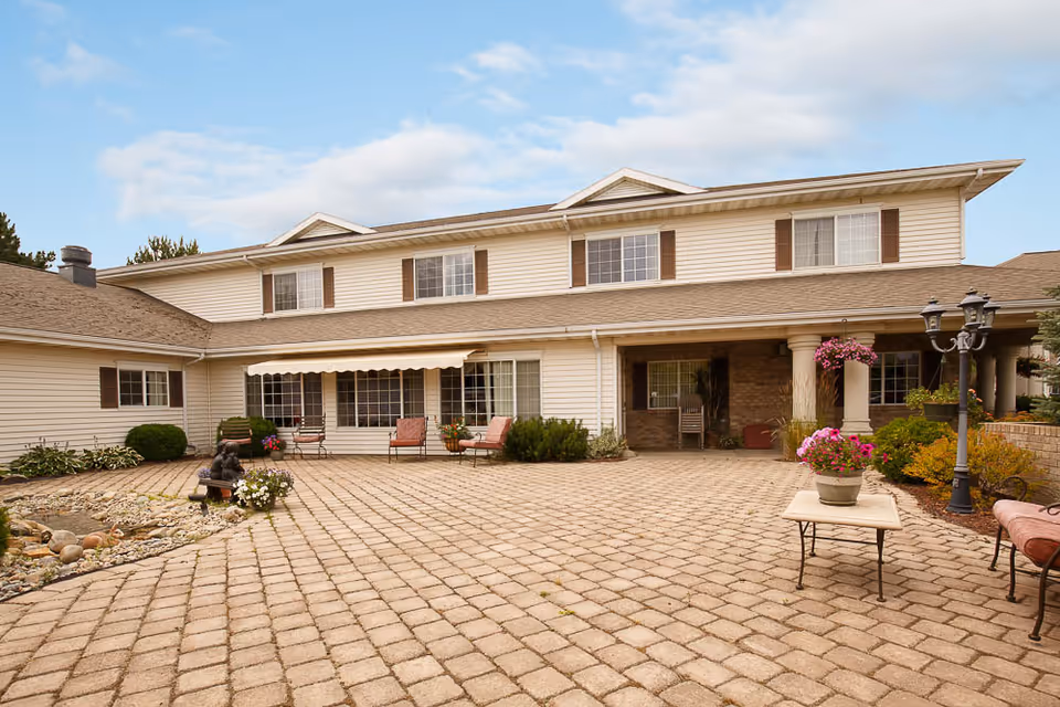 Two-story beige senior living building with a large paved courtyard, patio seating, potted flowers and a small rock garden.