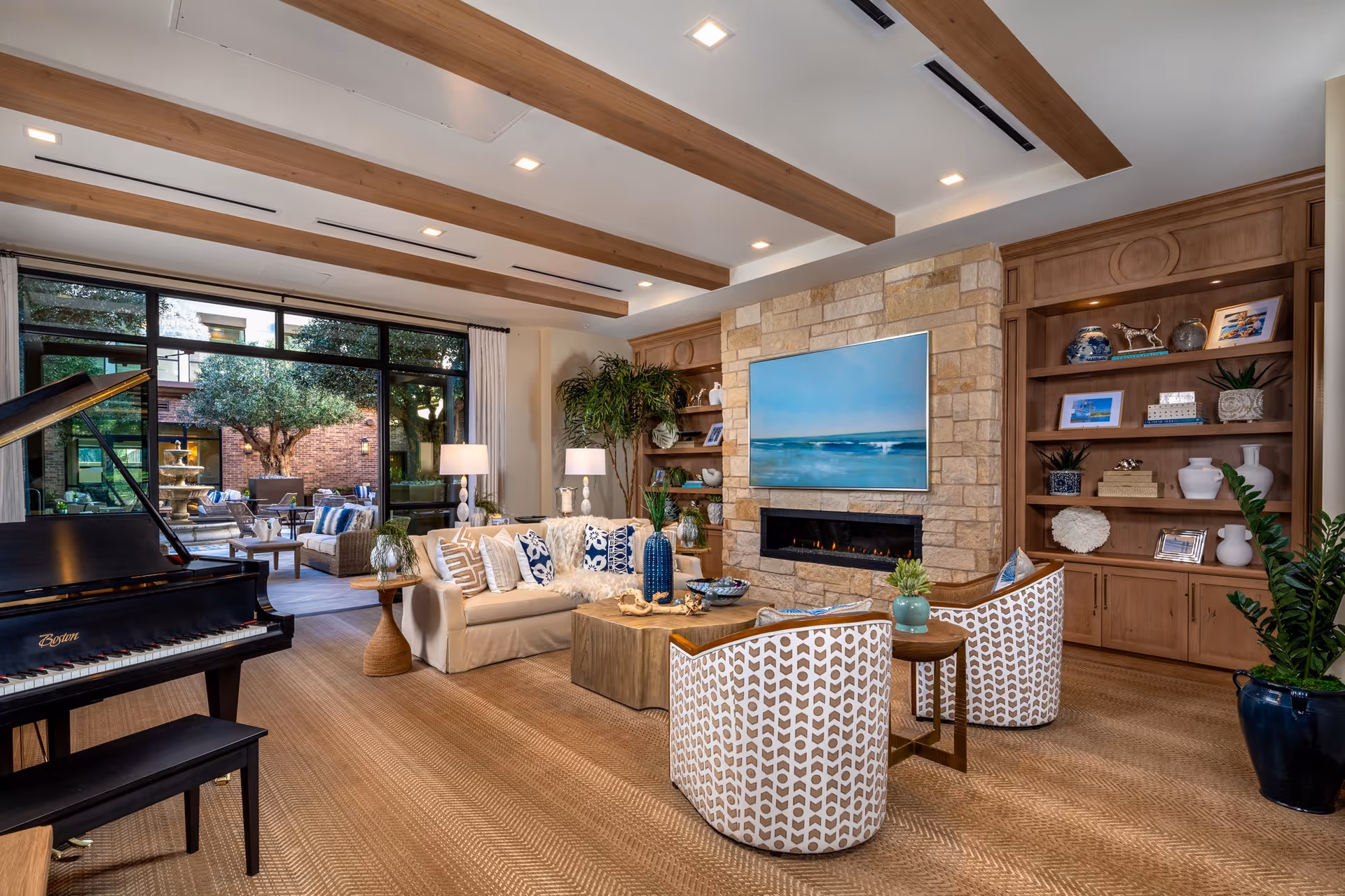 A cozy living room with a beige sofa adorned with patterned pillows, two patterned armchairs, a wooden coffee table, and a black grand piano. The room features a stone fireplace with a large flat-screen TV mounted above it, built-in wooden shelves with decorative items, and large windows showing an outdoor patio with seating and a fountain.