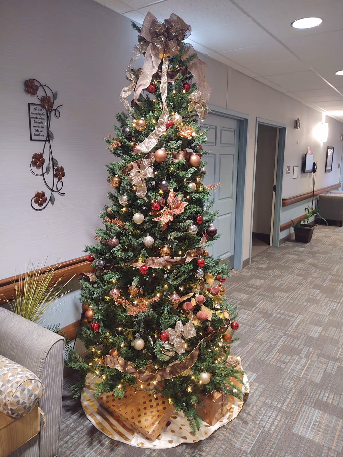 A decorated Christmas tree with gold, red, and bronze ornaments and ribbons stands in a hallway of a senior living facility. The tree is surrounded by wrapped presents at the base. The hallway has light-colored walls, carpeted floor, and several doors along the corridor. A cushioned chair and some wall decorations are visible nearby.
