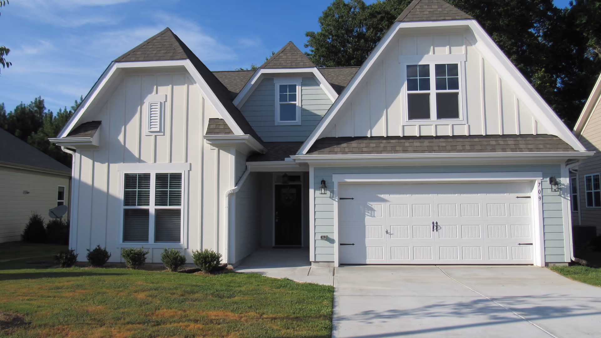 Front exterior view of a modern two-story house with white and light blue siding, a double garage door, and a small front porch. The house has multiple gabled roofs and several windows. There is a well-maintained lawn in front.