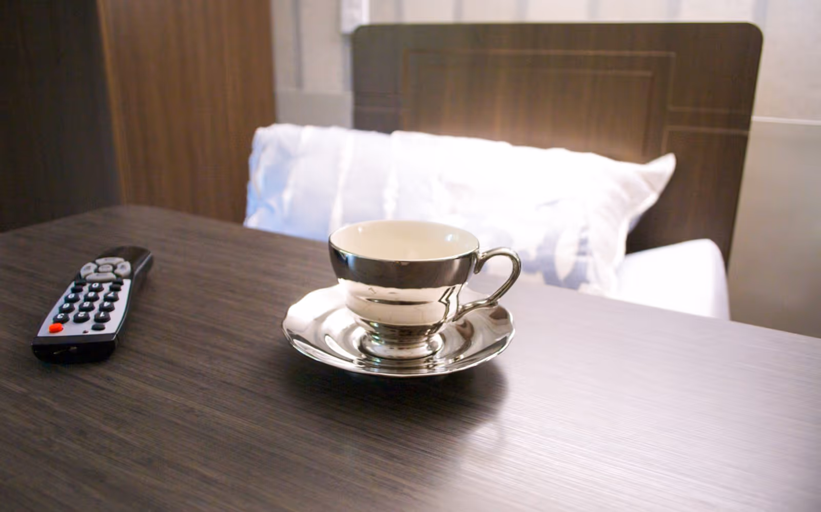 A close-up view of a dark wooden table with a silver cup and saucer placed on it. A TV remote control is also on the table. In the background, there is a bed with white pillows and a dark wooden headboard.