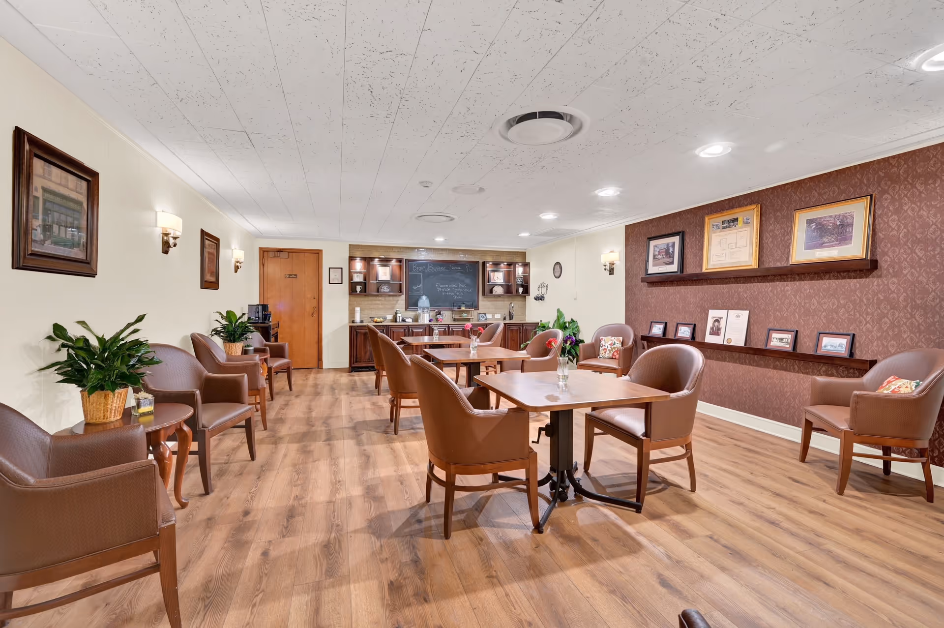 A well-lit common area in a senior living facility featuring wooden flooring, several brown leather chairs arranged around small wooden tables with flower vases, framed pictures on the walls, and a kitchenette area with cabinets and a chalkboard in the background.