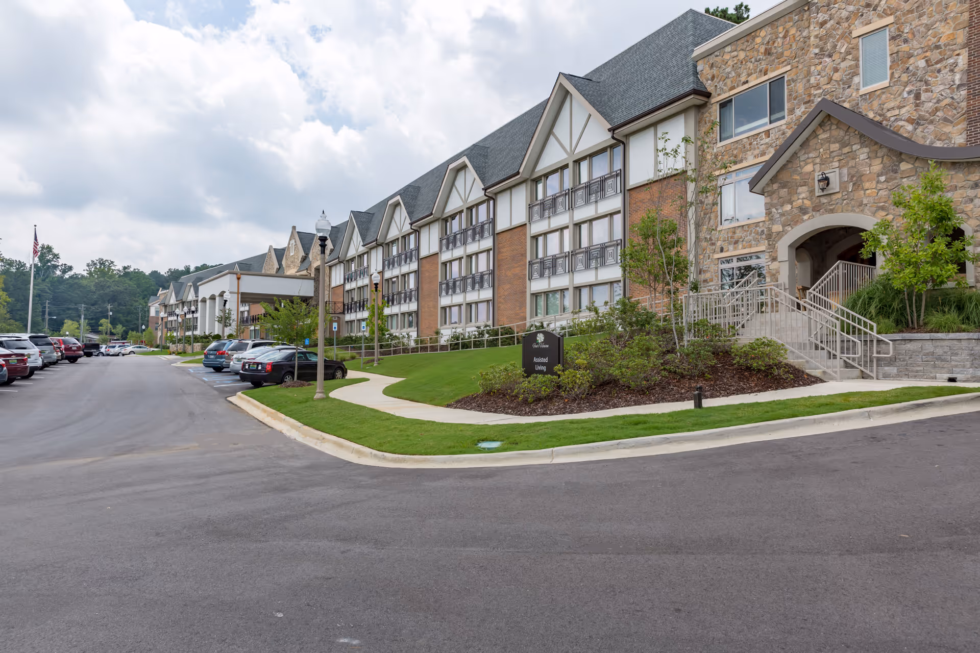 Exterior view of a large senior living facility building with stone and brick facade, multiple windows, and a sloped roof. There is a paved driveway and parking area with several cars parked. A sign near the entrance reads 'Assisted Living'. The sky is partly cloudy.