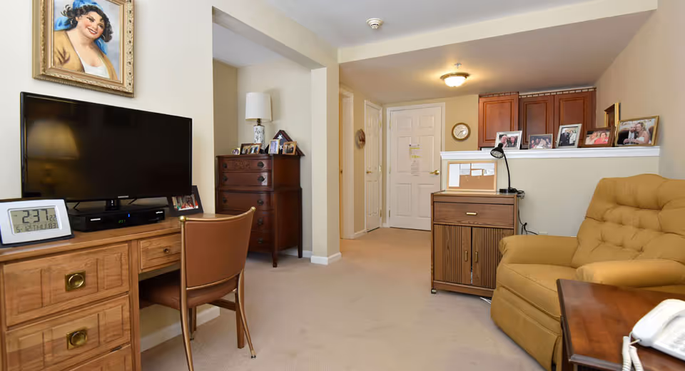 Interior view of a living area in an assisted living facility featuring a wooden desk with a flat-screen TV, a brown chair, a chest of drawers with a lamp and framed photos, a cushioned armchair, a small cabinet with a lamp and more framed photos, and a hallway leading to a white door.