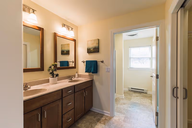A bathroom with a double sink vanity featuring wooden cabinets and two mirrors above. There are two wall-mounted light fixtures above each mirror. A small potted plant and a soap dispenser are on the countertop. A teal towel hangs on a towel rack on the wall, and a framed landscape painting is above the towel. The bathroom opens into a room with a window and tiled floor.