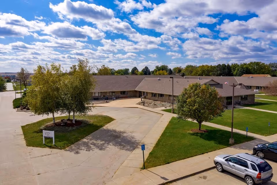 Aerial view of a single-story care facility with a driveway, parking lot, lawns and trees under a partly cloudy sky.