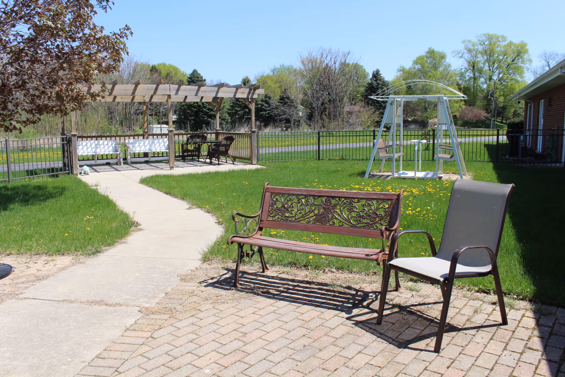 Outdoor seating area with a decorative metal bench and a cushioned chair on a paved patio. A concrete pathway leads to a wooden pergola with white benches and a table with chairs underneath. In the background, there is a swing set with two seats and a grassy area surrounded by a black metal fence. Trees and a clear blue sky are visible in the distance.