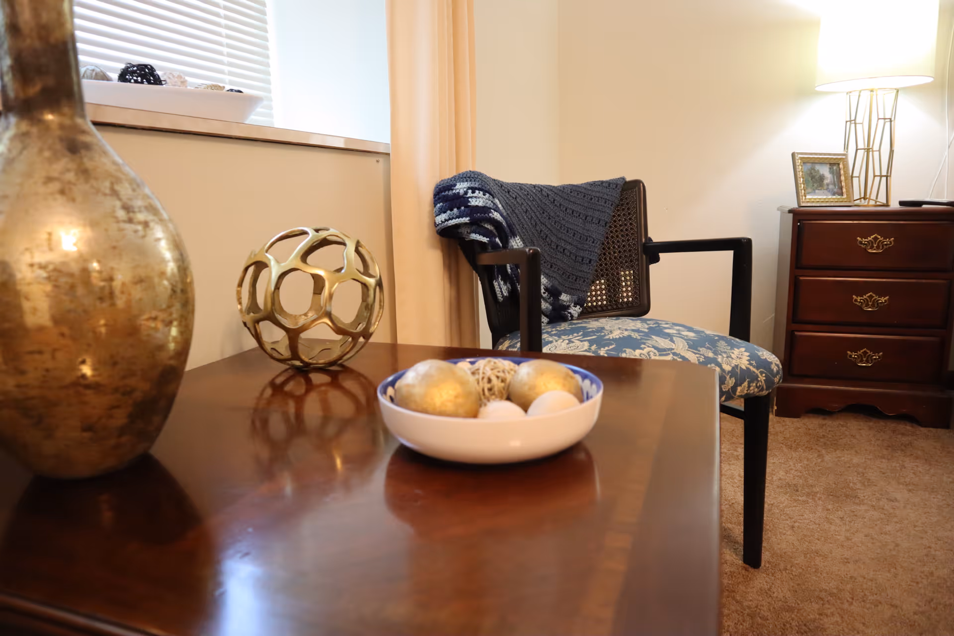 A cozy corner of a room featuring a wooden table with decorative items including a gold vase, a gold geometric sculpture, and a bowl with decorative balls. In the background, there is a cushioned chair with a blue patterned seat and a dark blue knitted throw draped over it. Next to the chair is a wooden nightstand with three drawers, a framed picture, and a lamp with a white shade. The room has beige walls and carpeted floor.