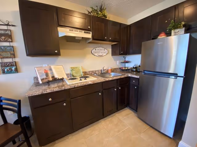 A compact kitchen with dark brown cabinets, a marble-patterned countertop, a stainless steel refrigerator, a small sink, and a stovetop. There is a 'Home Sweet Home' sign on the wall above the sink, a few decorative items on the counter, and a chair partially visible on the left side.