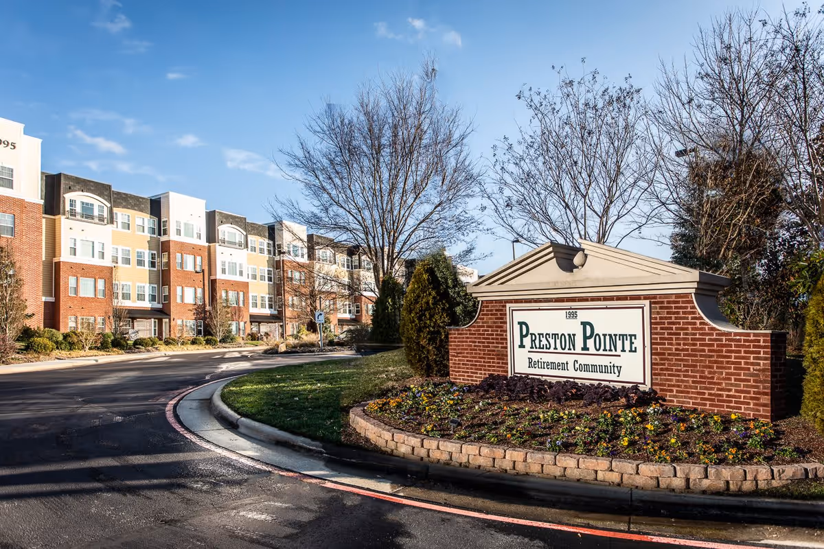 Exterior view of Preston Pointe Retirement Community showing a multi-story residential building with a brick and beige facade, a curved driveway, landscaped greenery, and a brick sign with the community name.