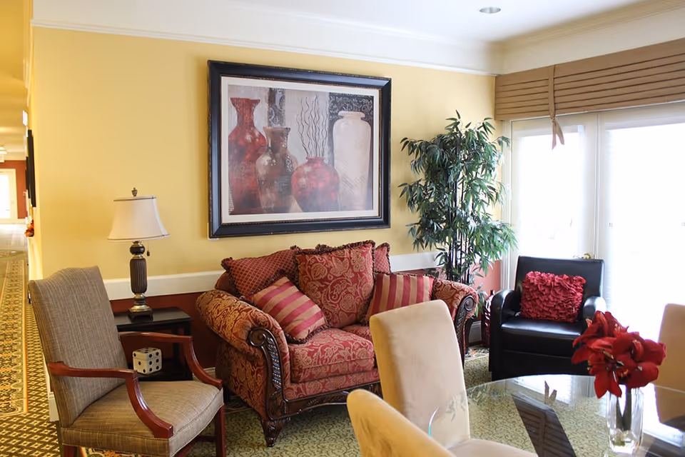 Cozy senior living lounge with a patterned sofa, armchairs, glass table, wall art and large windows.