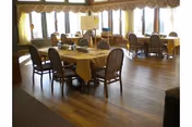 A spacious dining room with several wooden tables covered with yellow tablecloths and surrounded by wooden chairs. Large windows with curtains allow natural light to fill the room, and a television is mounted on a stand near the windows.