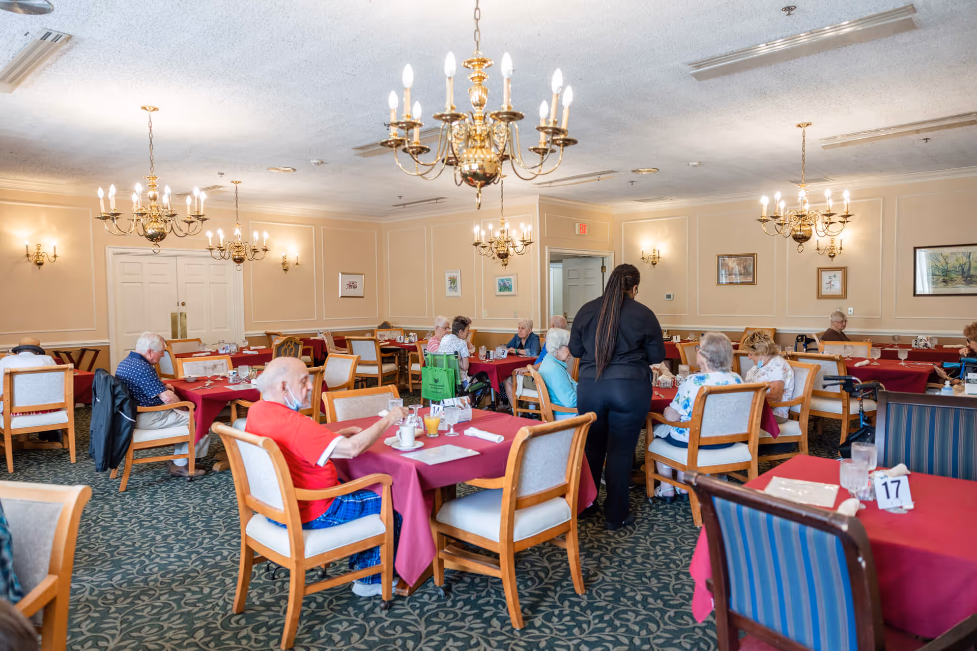 A dining room in a senior living facility with several elderly residents seated at tables covered with red tablecloths. A staff member is attending to some of the residents. The room features chandeliers, wall sconces, framed artwork, and a patterned carpet.