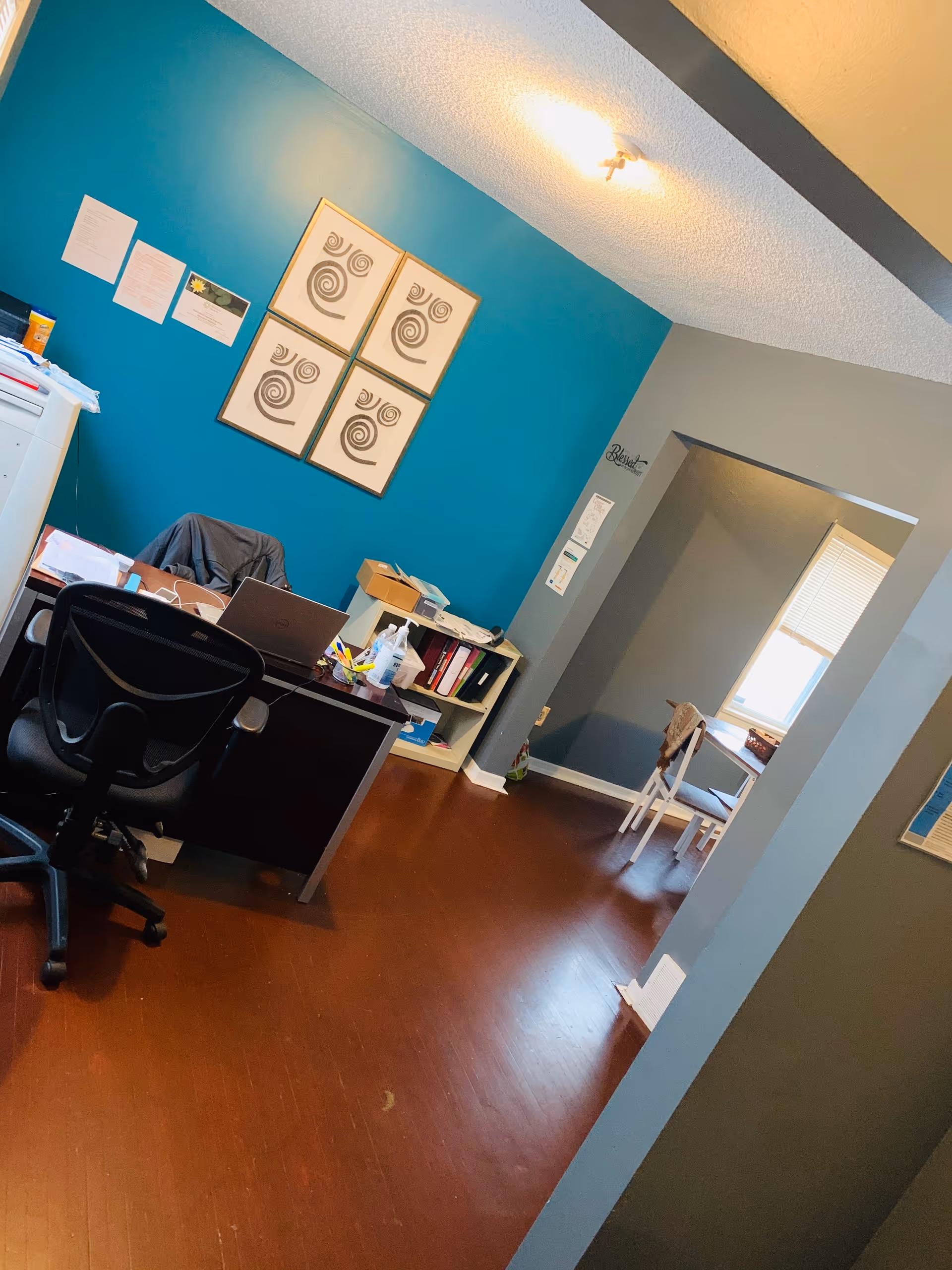 An office space with a black office chair and desk with a laptop, papers, and office supplies. The wall behind the desk is painted teal and has four framed abstract art pieces. There is a small shelf with binders and boxes next to the desk. The floor is wooden, and there is an open doorway leading to another room with a window and a chair.