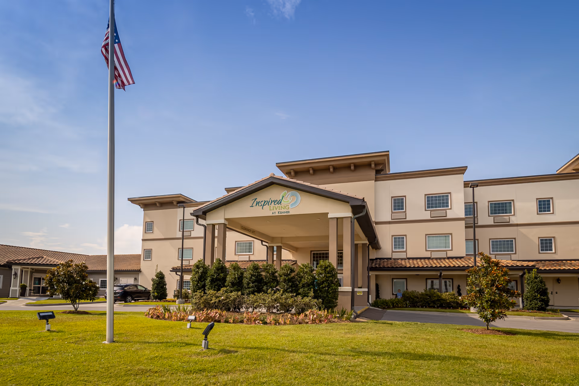 Exterior view of Inspired Living Kenner facility with a covered entrance, manicured bushes, a green lawn, and an American flag on a flagpole under a clear blue sky.
