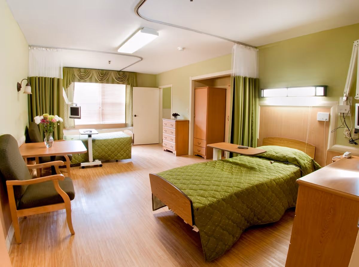 A well-lit senior living bedroom with two single beds covered in green quilts. The room features wooden furniture including a dresser, wardrobe, and bedside tables. There is a small table with a vase of flowers and two chairs near the window with green curtains. Medical equipment is mounted on the wall near one of the beds.