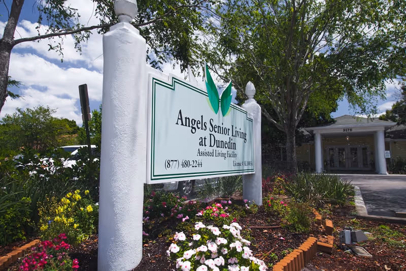Outdoor view of the entrance area of Angels Senior Living at Dunedin, featuring a white sign with green accents displaying the facility name and contact number, surrounded by colorful flowers and greenery, with the building entrance visible in the background under a partly cloudy sky.